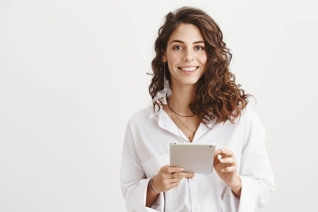 successful caucasian woman in trendy blouse holding tablet and smiling cheerfully at camera, expressing confidence while standing against gray background. girl in call centre ready to offer her help